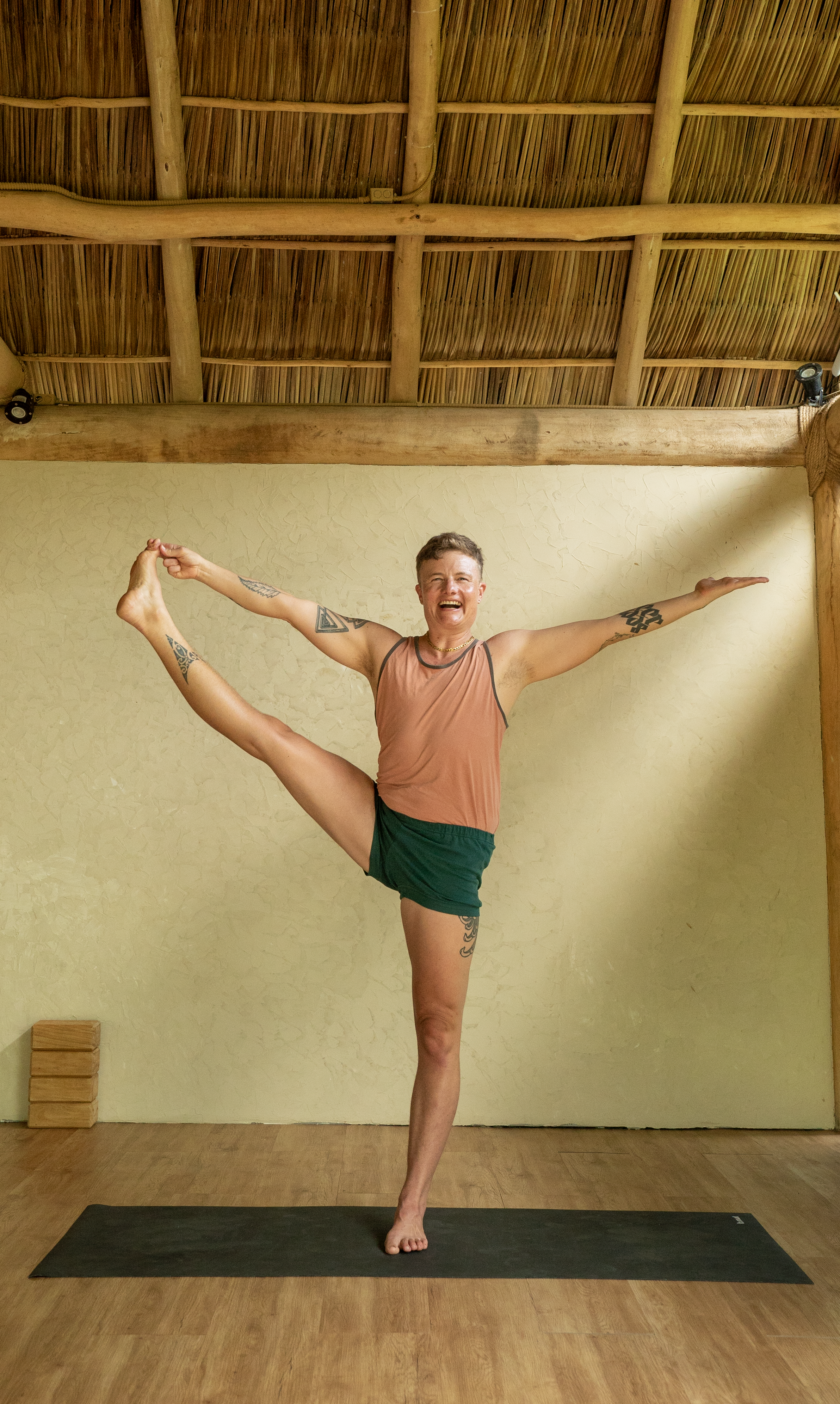 Avery Kalapa CIYT, a queer trans Iyengar Yoga teacher, smiling in an alignment based yoga asana standing on one foot with their arms extended wide.