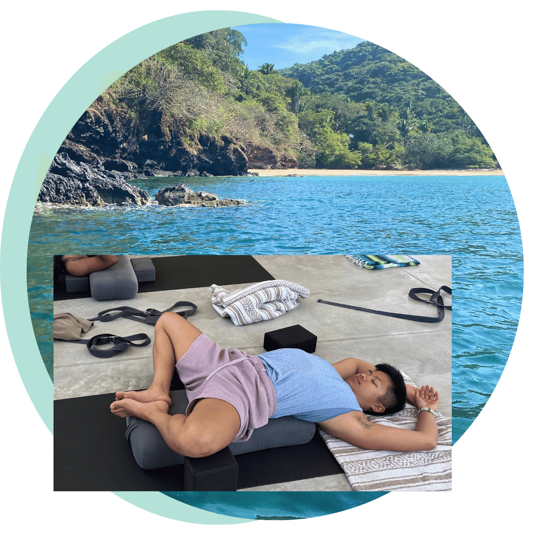 A queer Asian non-binary person relaxes in a restorative yoga pose with a picture of tropical ocean behind them.